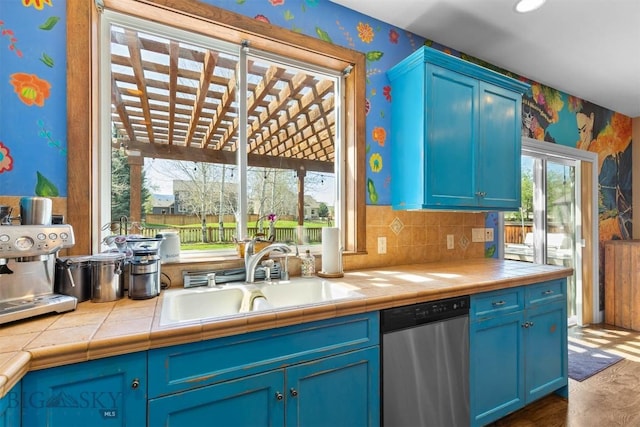 kitchen with wallpapered walls, a sink, stainless steel dishwasher, blue cabinets, and tile counters
