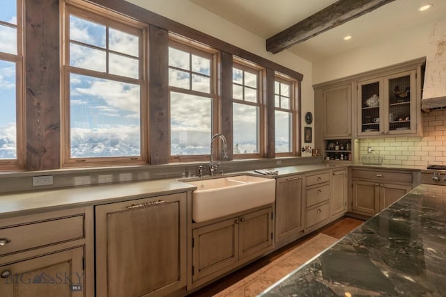 kitchen featuring a sink, decorative backsplash, beam ceiling, glass insert cabinets, and light wood-type flooring