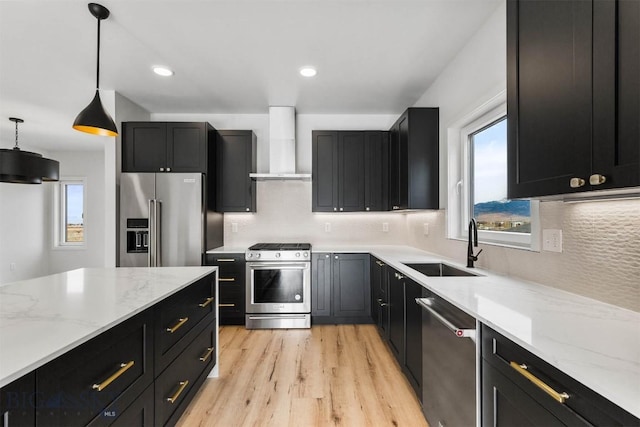 kitchen featuring appliances with stainless steel finishes, wall chimney range hood, a sink, dark cabinets, and light wood-type flooring