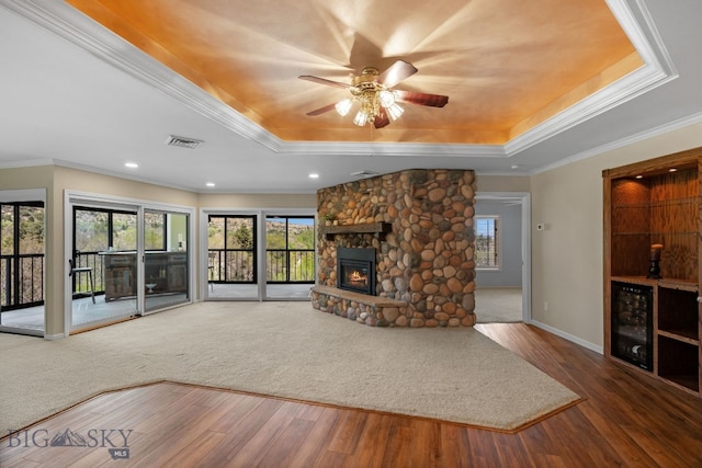 unfurnished living room featuring a tray ceiling, beverage cooler, a stone fireplace, wood finished floors, and ornamental molding