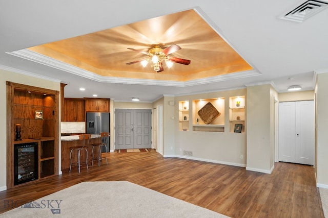 unfurnished living room with wine cooler, a raised ceiling, wood finished floors, a ceiling fan, and crown molding