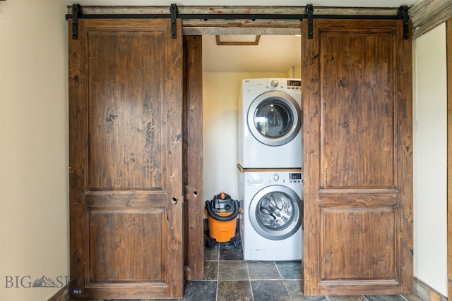 laundry area featuring stacked washer / drying machine, stone finish flooring, and a barn door