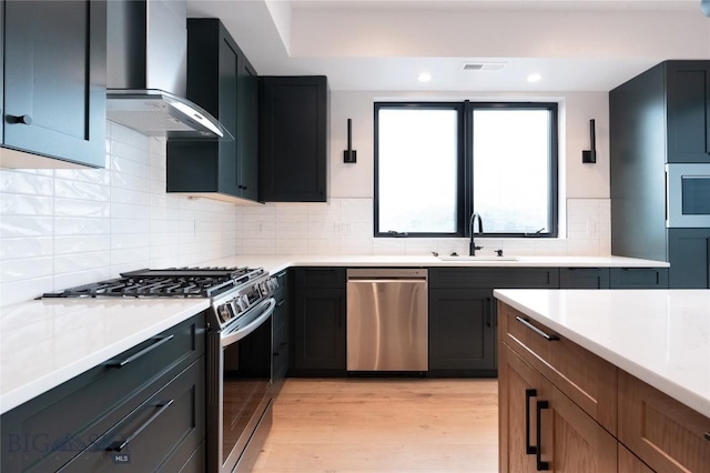 kitchen featuring appliances with stainless steel finishes, wall chimney range hood, a sink, light wood finished floors, and light countertops
