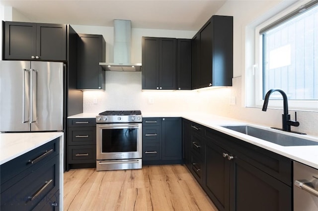 kitchen with stainless steel appliances, wall chimney range hood, a sink, light wood-style floors, and light countertops