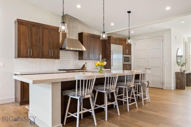 20 Caymus Ln Unit D, Bozeman, MT 59718 - kitchen with wall chimney range hood, stainless steel appliances, light wood-style flooring, a kitchen island with sink, and backsplash