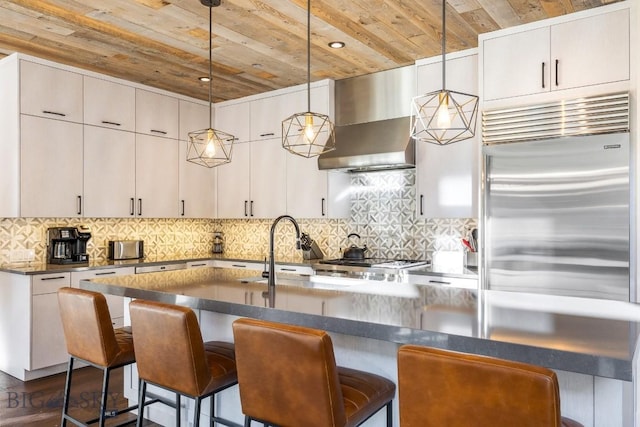 425 Outlook Trl, Big Sky, MT 59716 - kitchen with built in fridge, wall chimney range hood, wood ceiling, dark countertops, and a kitchen breakfast bar