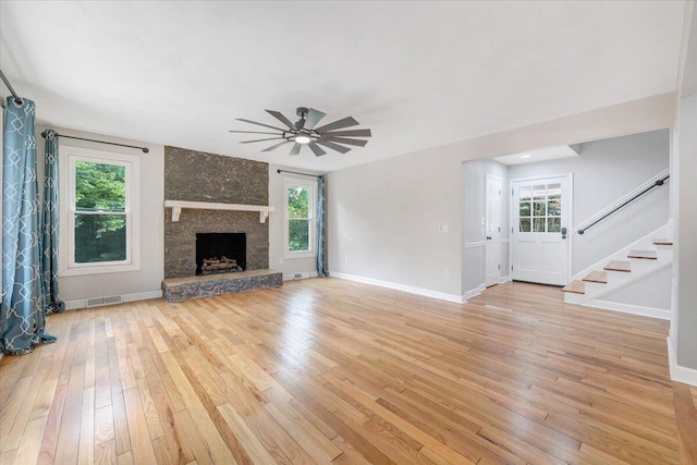 unfurnished living room featuring a stone fireplace, visible vents, a ceiling fan, stairway, and light wood-type flooring