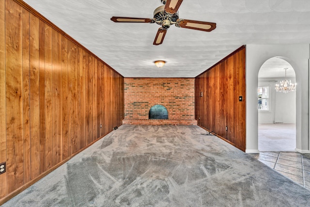 unfurnished living room with arched walkways, carpet floors, ceiling fan with notable chandelier, and wooden walls