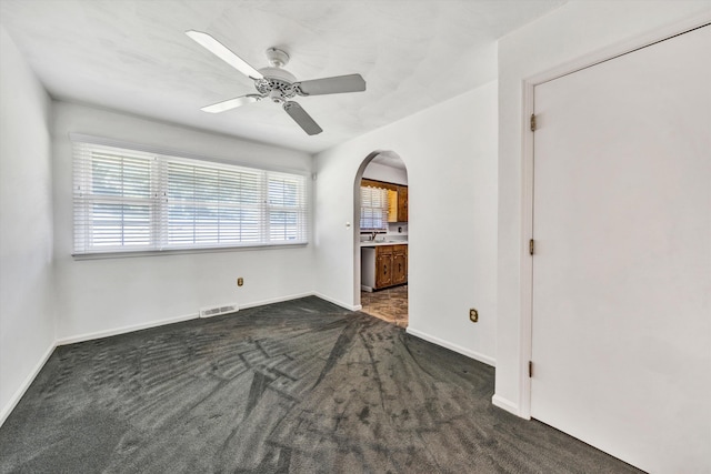 empty room with baseboards, visible vents, arched walkways, a ceiling fan, and dark colored carpet