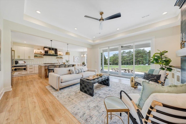 living area featuring visible vents, a tray ceiling, recessed lighting, light wood-style floors, and ceiling fan with notable chandelier