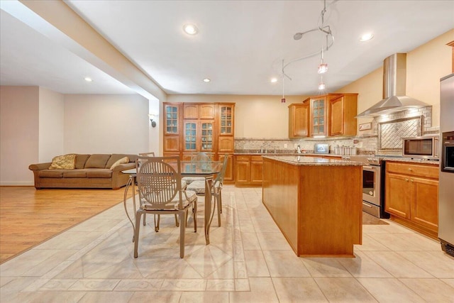 kitchen with wall chimney exhaust hood, light stone counters, tasteful backsplash, appliances with stainless steel finishes, and a kitchen island