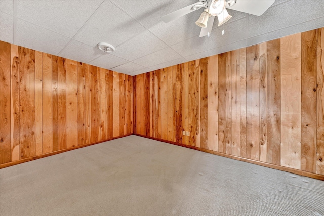 carpeted spare room featuring baseboards, a drop ceiling, a ceiling fan, and wooden walls