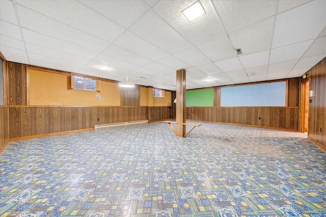 basement with a baseboard radiator, a drop ceiling, and wood walls