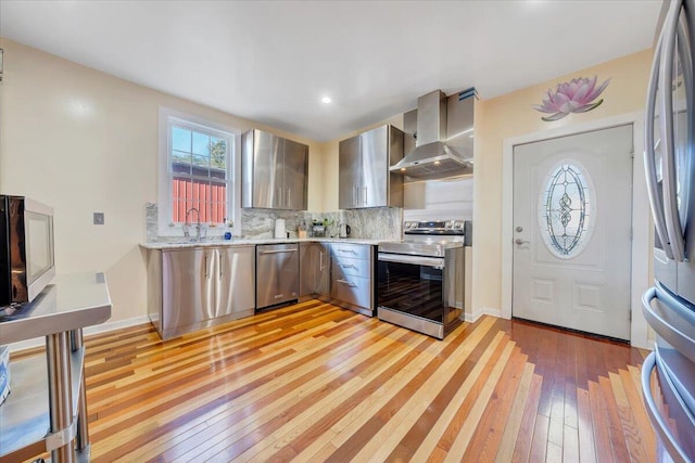 kitchen with sink, wall chimney range hood, stainless steel appliances, light hardwood / wood-style floors, and decorative backsplash