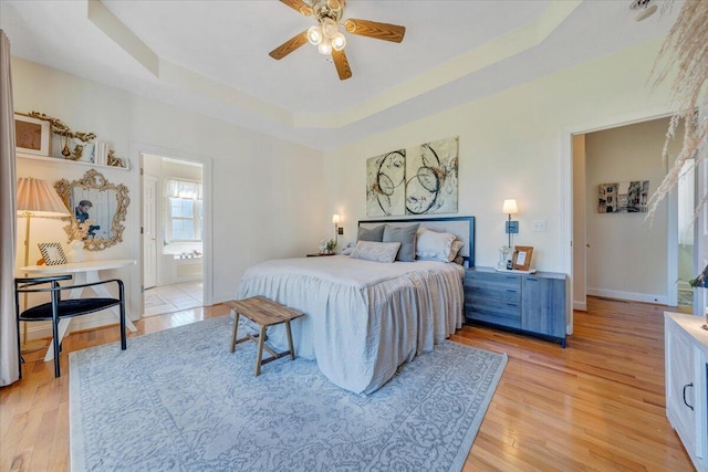 bedroom featuring ceiling fan, light wood-type flooring, ensuite bath, and a tray ceiling