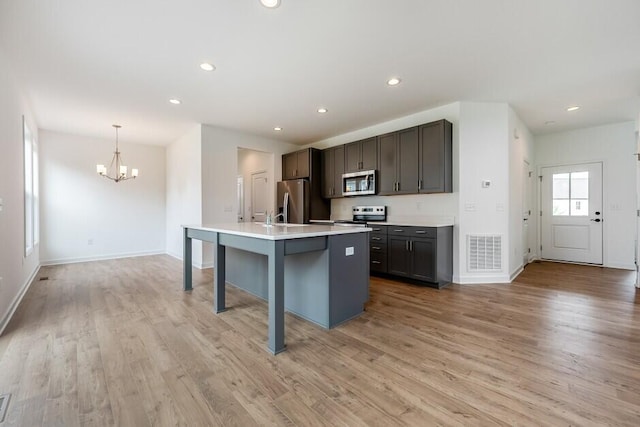 kitchen with an island with sink, an inviting chandelier, light wood-type flooring, stainless steel appliances, and decorative light fixtures