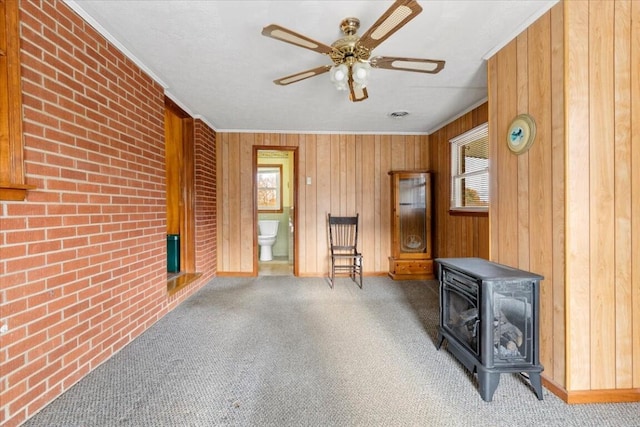 carpeted living room featuring brick wall, ceiling fan, wooden walls, crown molding, and a wood stove