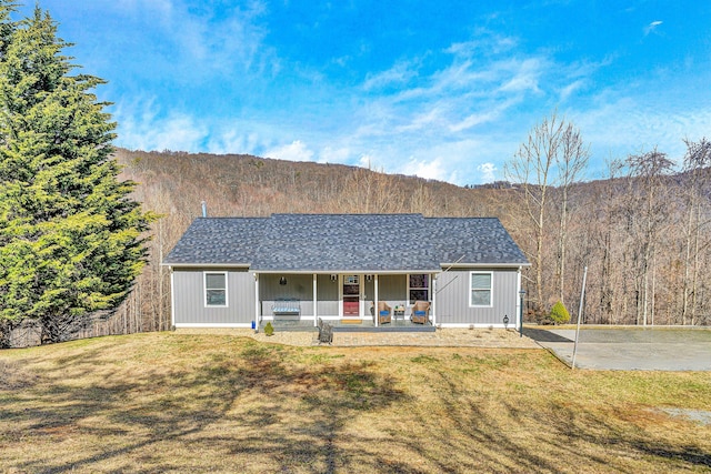 ranch-style home with covered porch, a view of trees, a front lawn, and a shingled roof