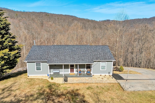 ranch-style house featuring covered porch, a view of trees, a front yard, and roof with shingles