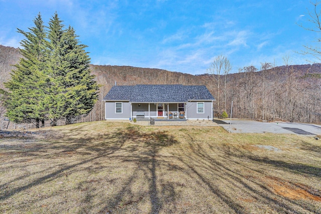 single story home featuring covered porch, a wooded view, a shingled roof, and a front yard