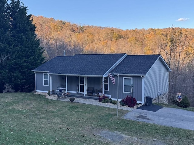 single story home featuring covered porch, a forest view, a front yard, and roof with shingles