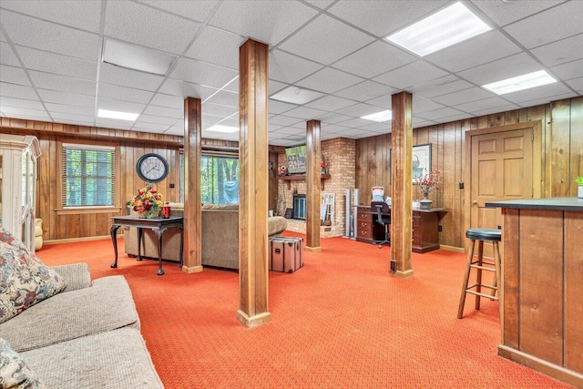 carpeted living room featuring a drop ceiling, a fireplace, wooden walls, and indoor bar