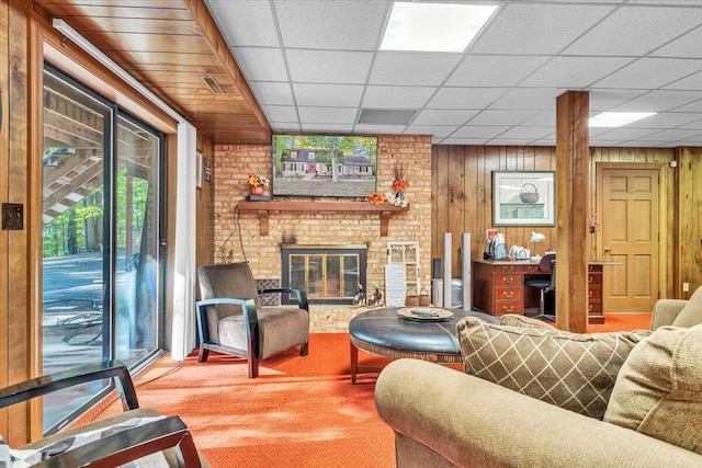 living room featuring a paneled ceiling, wood walls, carpet floors, and a brick fireplace