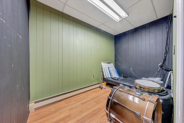 misc room featuring wood-type flooring, wooden walls, a drop ceiling, and a baseboard heating unit