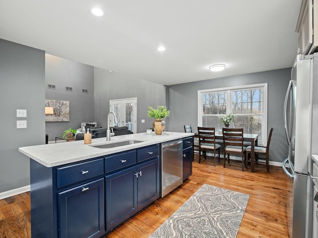 kitchen with sink, blue cabinets, an island with sink, appliances with stainless steel finishes, and light wood-type flooring