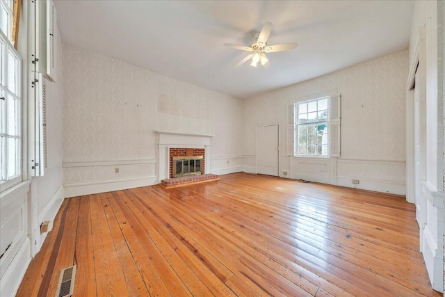 unfurnished living room featuring visible vents, a wainscoted wall, a ceiling fan, light wood-style floors, and a brick fireplace