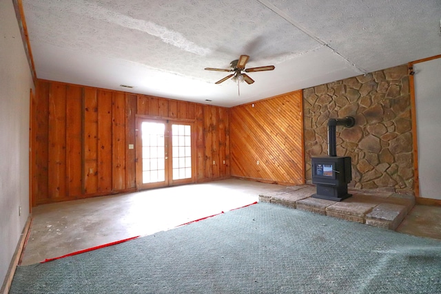 unfurnished living room with wood walls, a wood stove, ceiling fan, a textured ceiling, and french doors