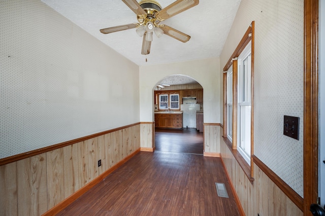 empty room with arched walkways, dark wood-style flooring, a wainscoted wall, visible vents, and wallpapered walls