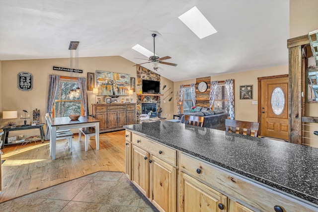 kitchen featuring ceiling fan, lofted ceiling with skylight, light brown cabinets, a stone fireplace, and light tile patterned floors