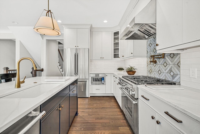 kitchen featuring wall chimney range hood, high end appliances, hanging light fixtures, a sink, and white cabinetry