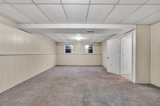 basement with a drop ceiling, wooden walls, and light colored carpet