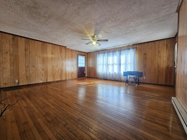 unfurnished room featuring dark hardwood / wood-style floors, wood walls, a baseboard heating unit, ceiling fan, and a textured ceiling