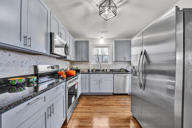 kitchen featuring appliances with stainless steel finishes, gray cabinets, pendant lighting, dark stone counters, and hardwood / wood-style floors