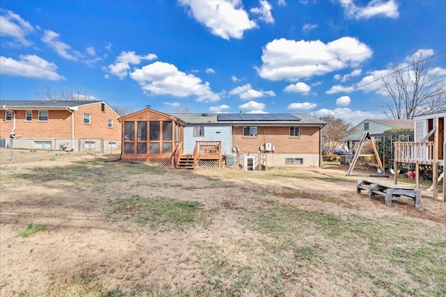 rear view of property featuring solar panels, a yard, a wooden deck, a playground, and a sunroom