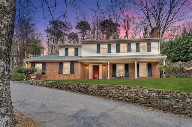 view of front of property featuring roof with shingles, a yard, covered porch, a chimney, and brick siding