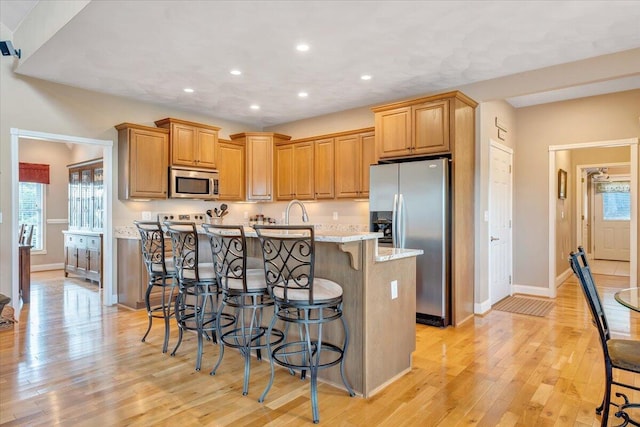 kitchen with a breakfast bar, appliances with stainless steel finishes, light hardwood / wood-style floors, light stone countertops, and a center island with sink