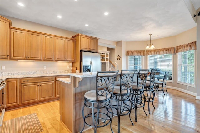 kitchen with stainless steel refrigerator with ice dispenser, decorative light fixtures, a kitchen island with sink, light stone countertops, and light hardwood / wood-style floors