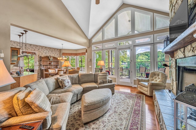 living room with high vaulted ceiling, a stone fireplace, ceiling fan with notable chandelier, wallpapered walls, and wood finished floors