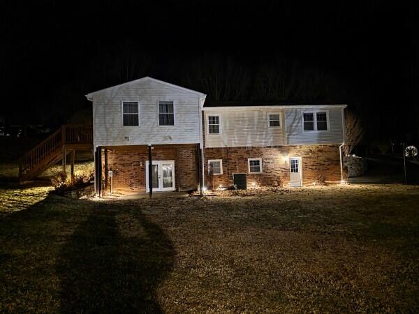 back of house at night featuring stairs, stone siding, a lawn, and central air condition unit