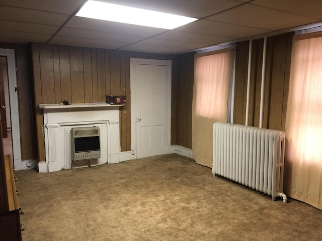 unfurnished living room with a drop ceiling, carpet floors, heating unit, wooden walls, and radiator