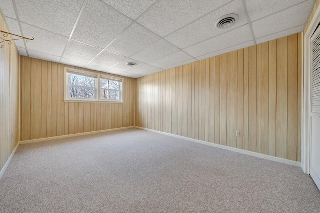 carpeted empty room with wooden walls, baseboards, a paneled ceiling, and visible vents