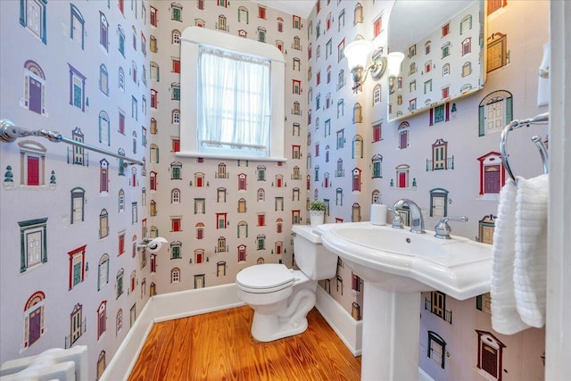 half bathroom featuring wallpapered walls, baseboards, toilet, wood finished floors, and a workshop area