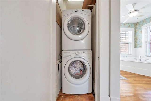 clothes washing area with laundry area, wood finished floors, a ceiling fan, stacked washer / drying machine, and wallpapered walls