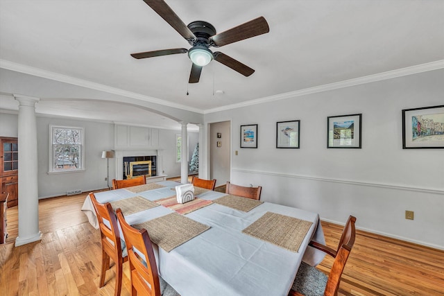 dining space featuring crown molding, a glass covered fireplace, light wood finished floors, ornate columns, and a ceiling fan