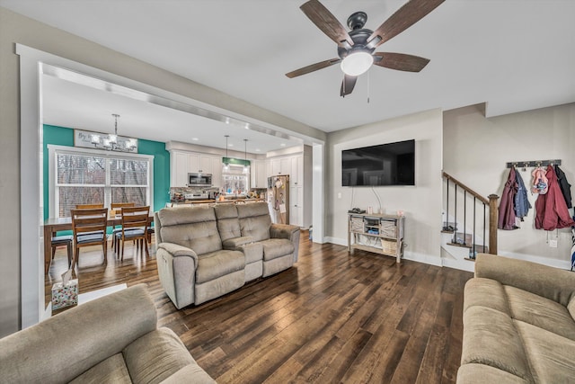 living room featuring stairs, dark wood-style flooring, ceiling fan with notable chandelier, and baseboards