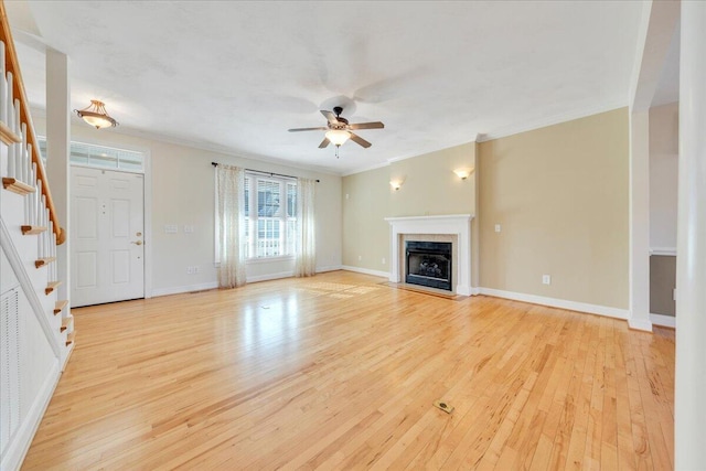 unfurnished living room featuring crown molding, light wood-style flooring, a fireplace with flush hearth, a ceiling fan, and stairs